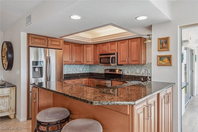 kitchen with appliances with stainless steel finishes, a sink, a tray ceiling, a peninsula, and brown cabinets