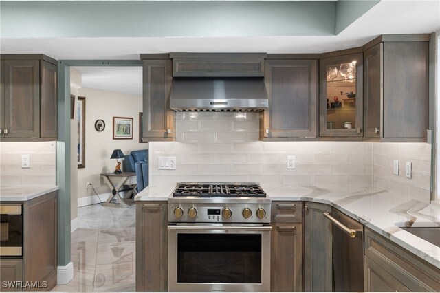 kitchen featuring light stone counters, stainless steel appliances, wall chimney range hood, tasteful backsplash, and glass insert cabinets