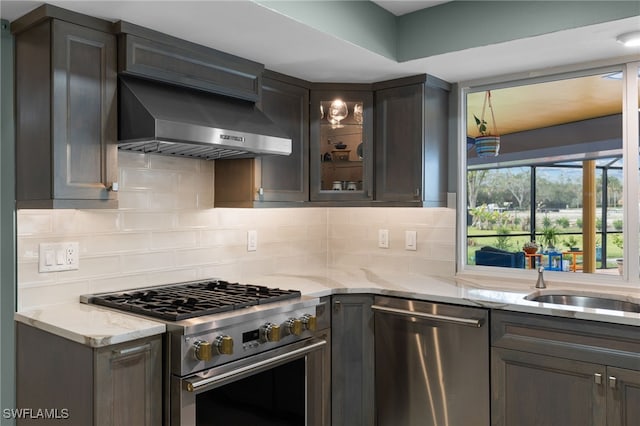 kitchen featuring light stone counters, stainless steel appliances, wall chimney range hood, decorative backsplash, and glass insert cabinets