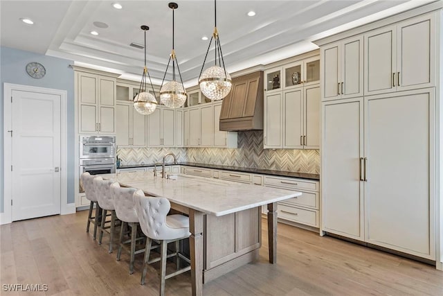 kitchen with custom exhaust hood, light wood-style flooring, double oven, and a raised ceiling