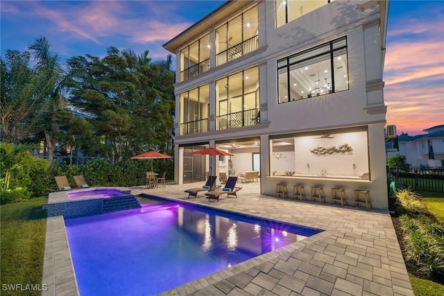 back of house at dusk featuring a patio, a balcony, fence, a pool with connected hot tub, and outdoor dry bar