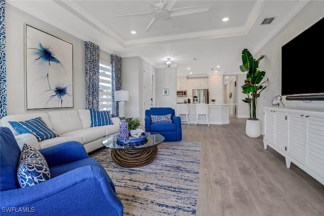 living room featuring ceiling fan, light wood-style flooring, visible vents, a tray ceiling, and crown molding