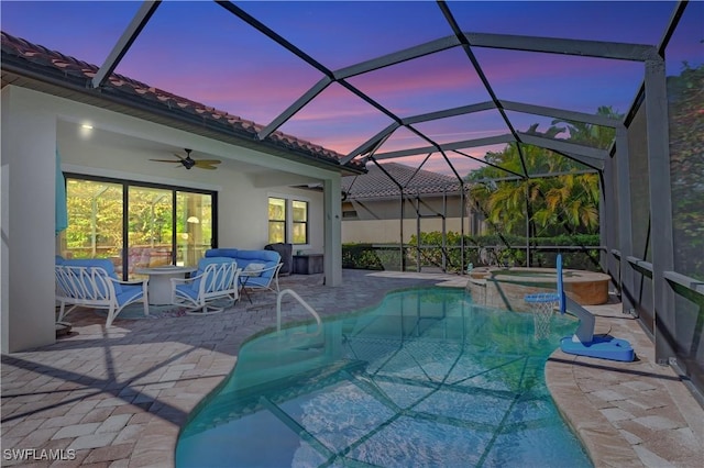 pool at dusk with a pool with connected hot tub, an outdoor hangout area, a ceiling fan, a patio area, and glass enclosure