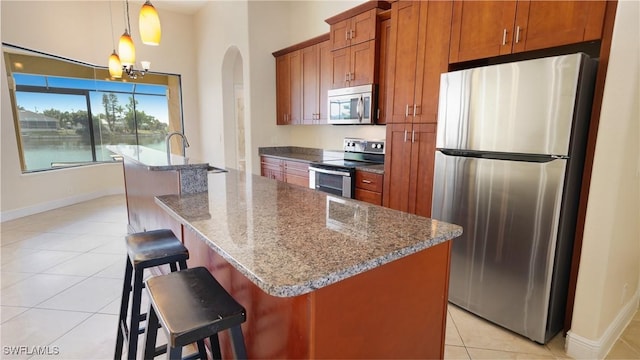 kitchen with a center island with sink, hanging light fixtures, stainless steel appliances, dark stone counters, and light tile patterned floors