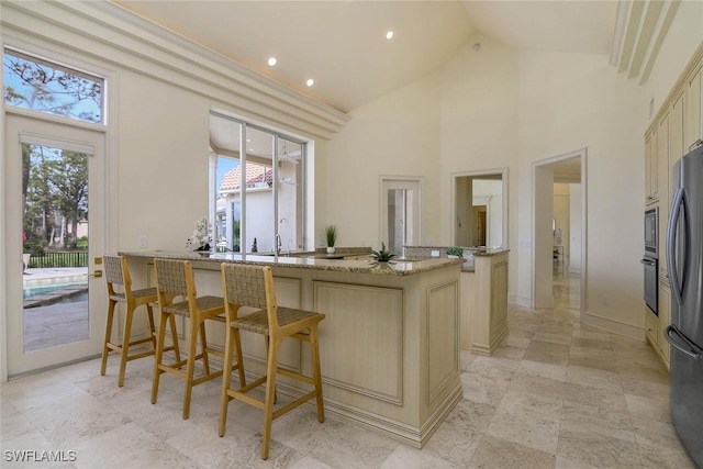 kitchen featuring a breakfast bar, light stone countertops, cream cabinets, stainless steel appliances, and high vaulted ceiling