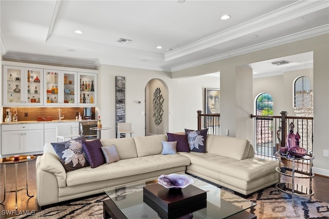 living area featuring arched walkways, wood finished floors, indoor wet bar, ornamental molding, and a tray ceiling