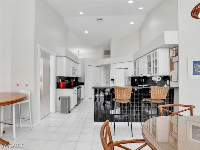 kitchen with glass insert cabinets, white fridge with ice dispenser, a peninsula, a kitchen bar, and white cabinetry