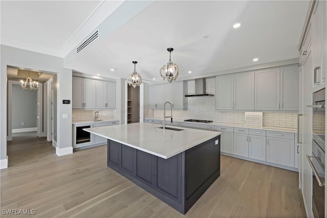 kitchen with a center island with sink, sink, pendant lighting, gray cabinetry, and wall chimney exhaust hood