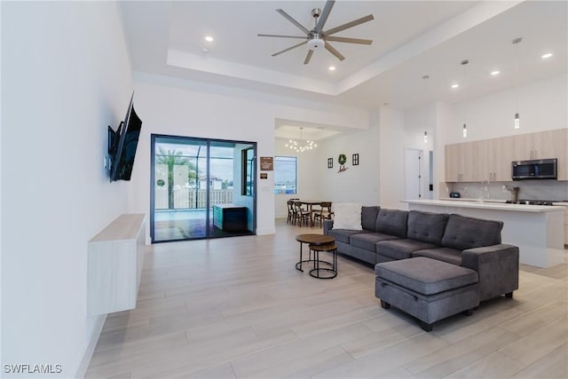 living area featuring recessed lighting, ceiling fan with notable chandelier, a high ceiling, light wood-type flooring, and a raised ceiling