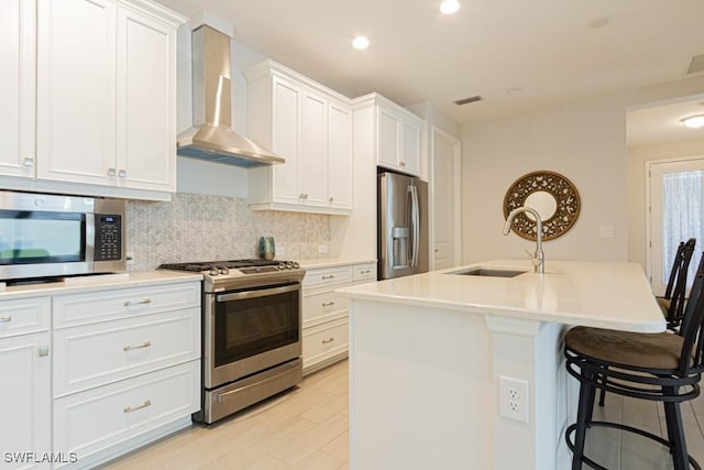 kitchen with appliances with stainless steel finishes, wall chimney exhaust hood, white cabinets, sink, and a kitchen island with sink