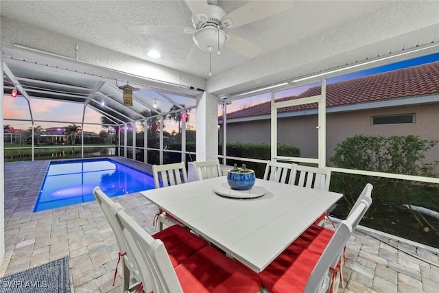 pool at dusk with glass enclosure, a patio, outdoor dining space, an outdoor pool, and ceiling fan