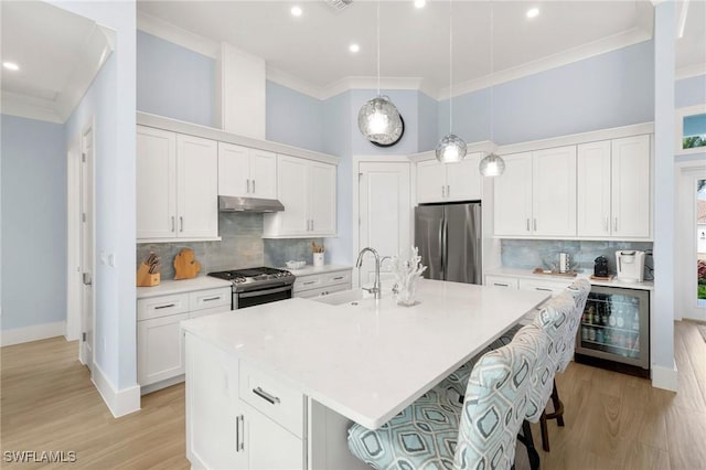 kitchen with beverage cooler, a center island with sink, appliances with stainless steel finishes, under cabinet range hood, and a sink