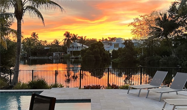 pool at dusk featuring a water view, fence, an outdoor pool, and a patio