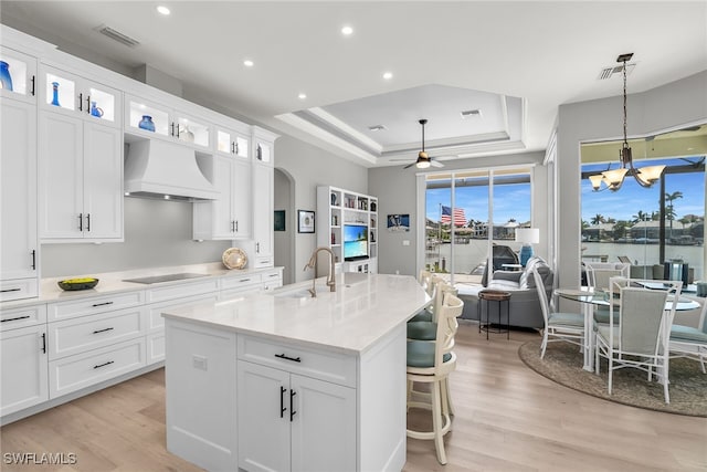 kitchen featuring a center island with sink, custom range hood, glass insert cabinets, decorative light fixtures, and white cabinetry