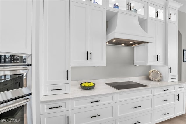 kitchen featuring light stone counters, custom exhaust hood, glass insert cabinets, stainless steel double oven, and white cabinetry