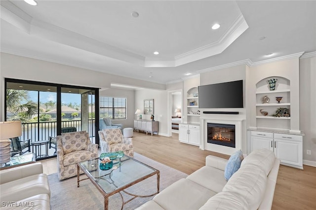 living area with built in shelves, a tray ceiling, crown molding, and light wood-style flooring