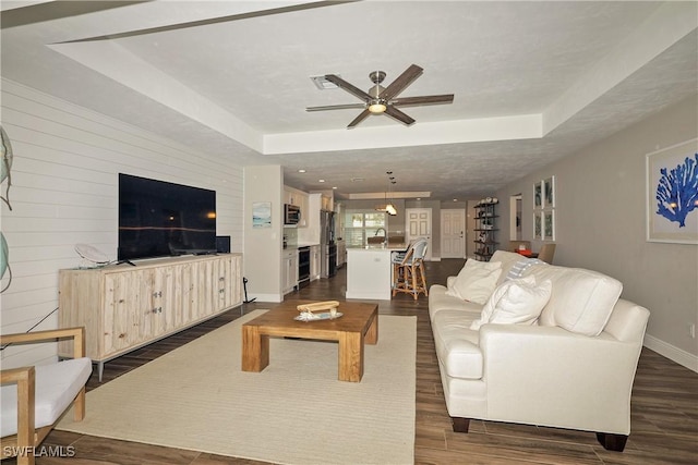 living room featuring baseboards, visible vents, a ceiling fan, dark wood-type flooring, and a tray ceiling