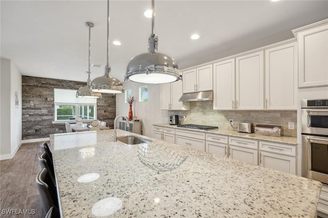 kitchen featuring light stone counters, under cabinet range hood, a sink, white cabinets, and decorative light fixtures