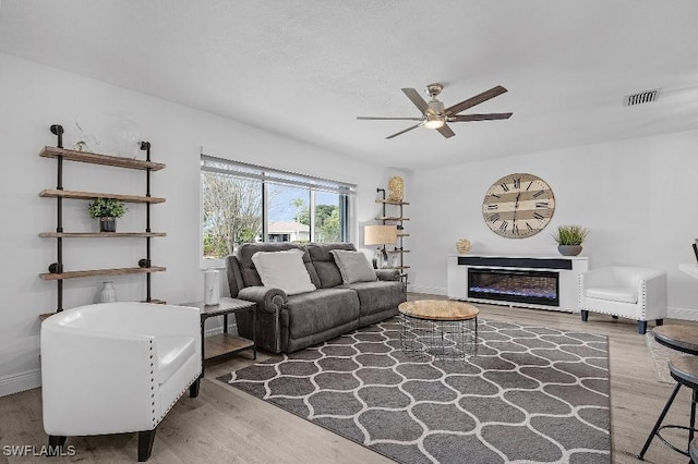 living area with wood finished floors, visible vents, a ceiling fan, baseboards, and a glass covered fireplace