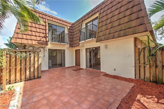 back of house with mansard roof, a patio, a balcony, fence, and stucco siding