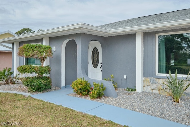 view of exterior entry featuring a shingled roof, a yard, and stucco siding