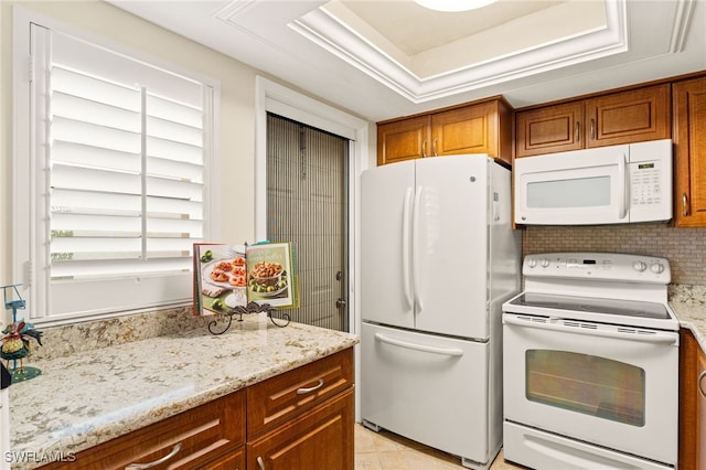 kitchen with white appliances, brown cabinets, light stone countertops, a tray ceiling, and backsplash