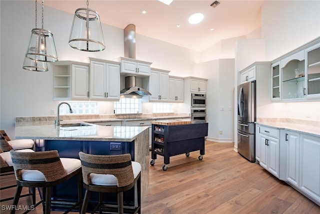 kitchen featuring stainless steel appliances, glass insert cabinets, a sink, a peninsula, and wall chimney exhaust hood