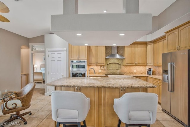 kitchen with appliances with stainless steel finishes, a center island with sink, wall chimney range hood, and light stone countertops