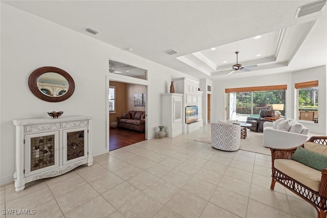 living room featuring light tile patterned floors, visible vents, a raised ceiling, a ceiling fan, and a glass covered fireplace