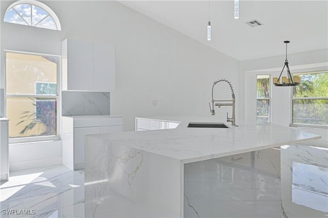 kitchen with hanging light fixtures, light stone counters, white cabinets, and an island with sink