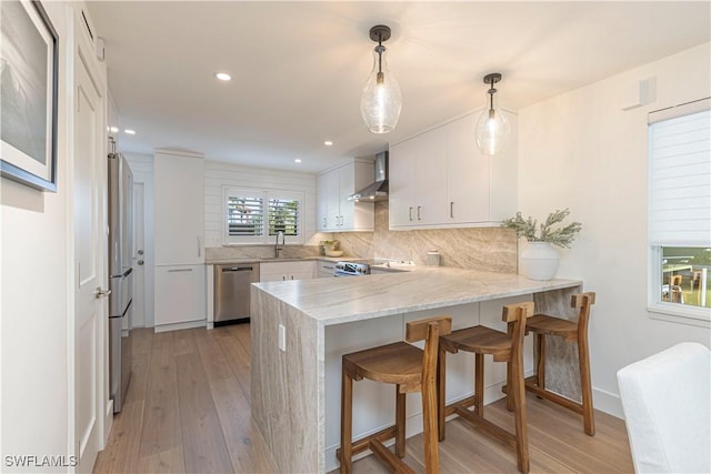 kitchen with stainless steel appliances, a peninsula, light wood-style floors, white cabinets, and wall chimney range hood