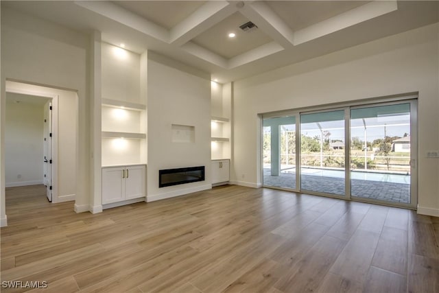 unfurnished living room featuring light wood finished floors, visible vents, coffered ceiling, a glass covered fireplace, and a high ceiling