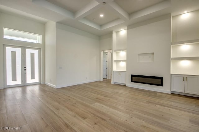 unfurnished living room featuring light wood-style flooring, a towering ceiling, a glass covered fireplace, coffered ceiling, and baseboards