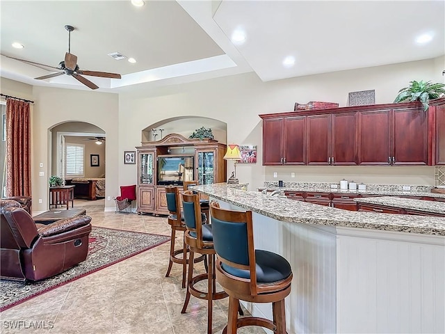 kitchen featuring arched walkways, light stone counters, a ceiling fan, open floor plan, and dark brown cabinets