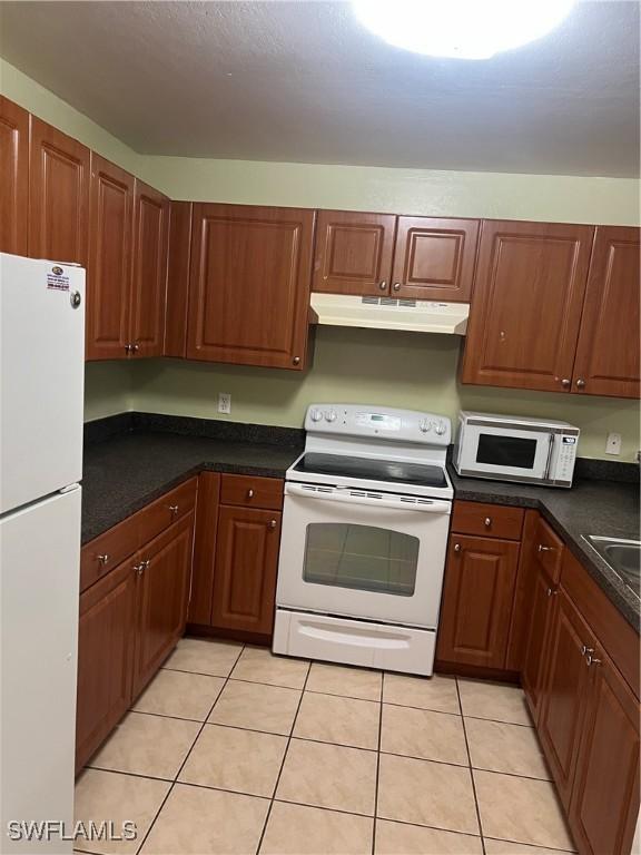 kitchen with light tile patterned floors, white appliances, dark countertops, and under cabinet range hood