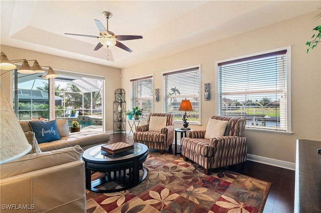 living room featuring dark wood-style flooring, a raised ceiling, a ceiling fan, a sunroom, and baseboards