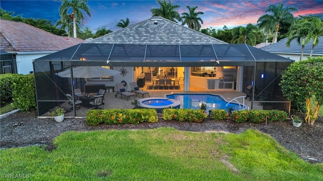 back of house at dusk featuring glass enclosure, a pool with connected hot tub, a patio area, and a lawn