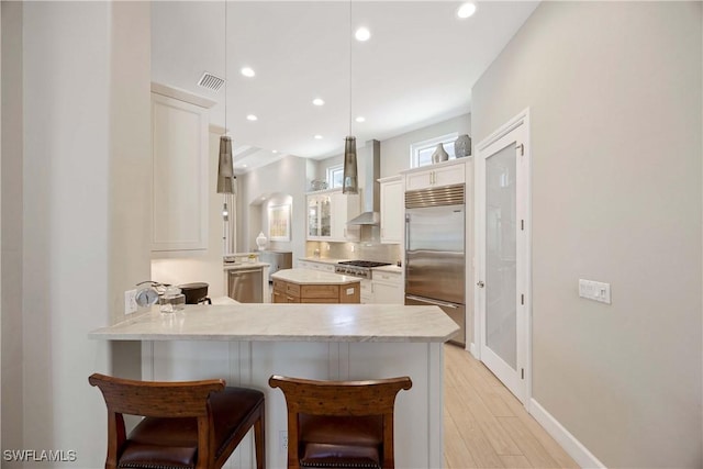 kitchen featuring visible vents, a peninsula, stainless steel built in fridge, light countertops, and wall chimney range hood