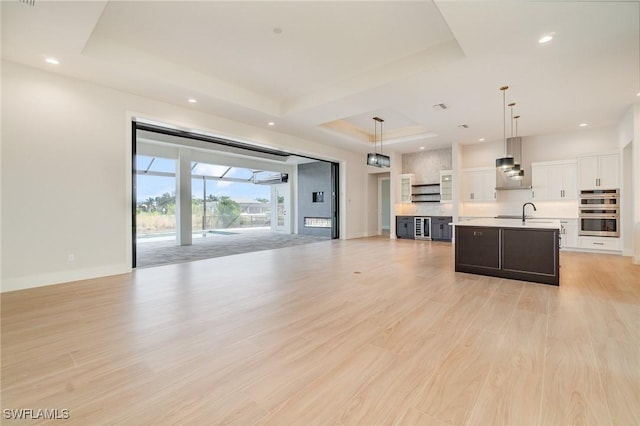 unfurnished living room featuring baseboards, light wood-style flooring, a tray ceiling, a sink, and recessed lighting