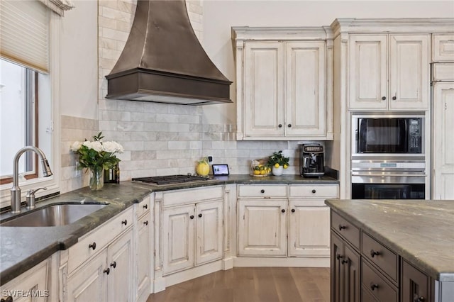 kitchen with dark stone countertops, a sink, custom exhaust hood, stainless steel appliances, and backsplash