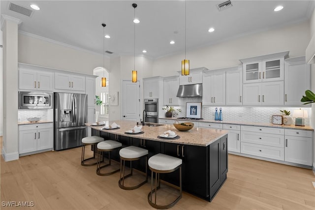 kitchen featuring under cabinet range hood, visible vents, appliances with stainless steel finishes, an island with sink, and crown molding