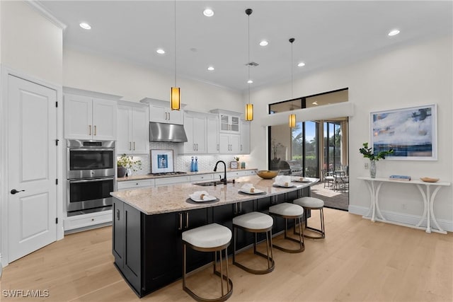 kitchen with light wood-style flooring, under cabinet range hood, double oven, white cabinetry, and backsplash