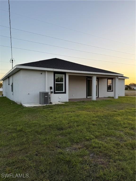 back of property at dusk with stucco siding, a shingled roof, central AC unit, and a yard