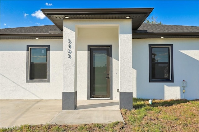 entrance to property featuring a shingled roof, a patio, a lawn, and stucco siding