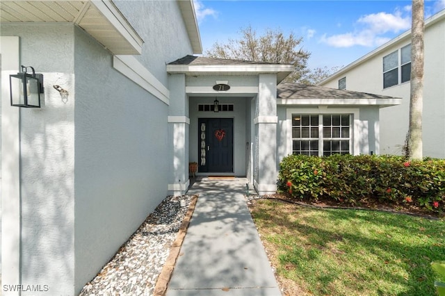 property entrance featuring roof with shingles, a lawn, and stucco siding