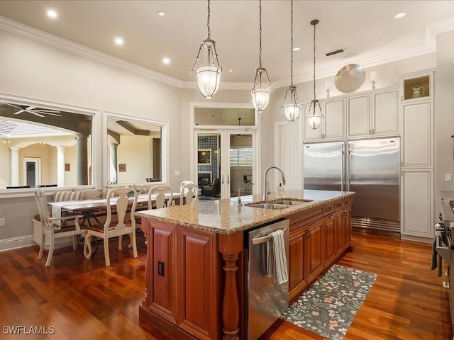 kitchen featuring a sink, appliances with stainless steel finishes, brown cabinetry, a center island with sink, and ornate columns