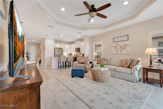 living area featuring ceiling fan, light wood-style flooring, recessed lighting, ornamental molding, and a tray ceiling