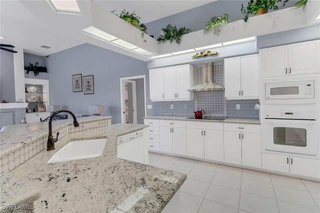 kitchen with white cabinetry, a sink, wall chimney range hood, light stone countertops, and white appliances