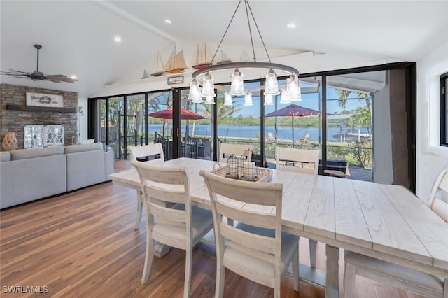 dining room featuring a water view, lofted ceiling with beams, ceiling fan, a stone fireplace, and wood finished floors