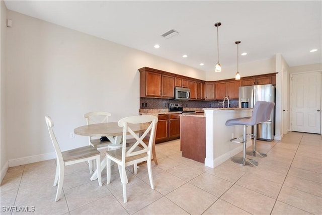 kitchen with a breakfast bar area, hanging light fixtures, appliances with stainless steel finishes, brown cabinetry, and an island with sink